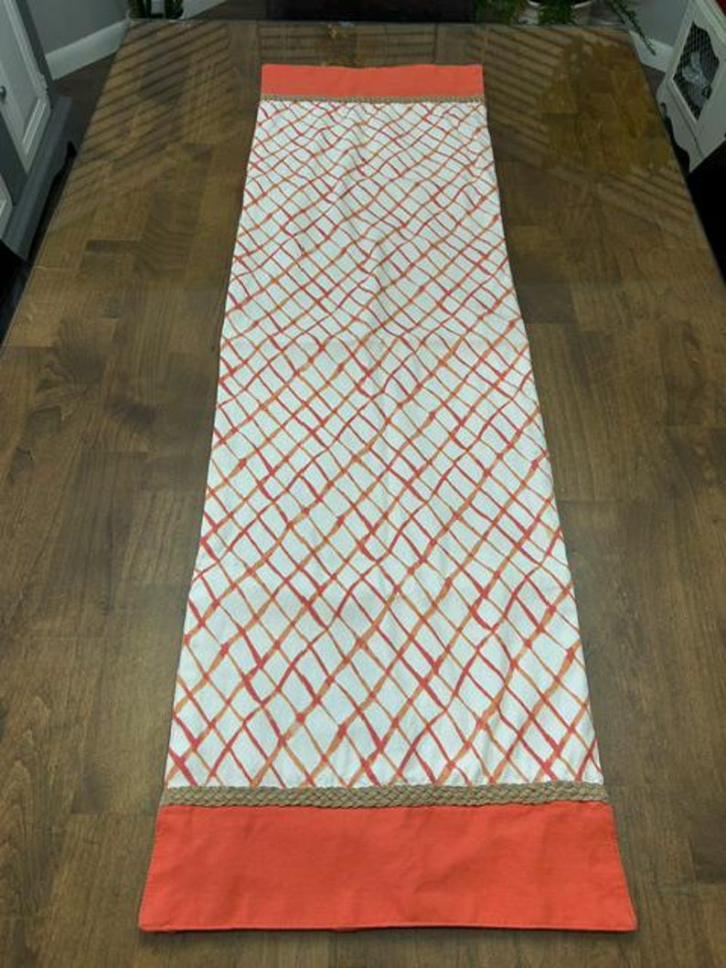 Puede incluir: Un camino de mesa rectangular con un panel central blanco que presenta un patr&oacute;n de cuadr&iacute;cula cruzada en naranja y rojo. El camino est&aacute; bordeado en ambos extremos por un ribete trenzado de color tostado y una banda de tela naranja s&oacute;lida.
