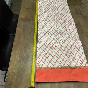 Puede incluir: Un camino de mesa rectangular con un centro blanco de patr&oacute;n de cuadr&iacute;cula naranja, bordeado por extremos de color naranja s&oacute;lido y ribete de yute trenzado. Una cinta m&eacute;trica amarilla al lado muestra una longitud de unos 117 cm.