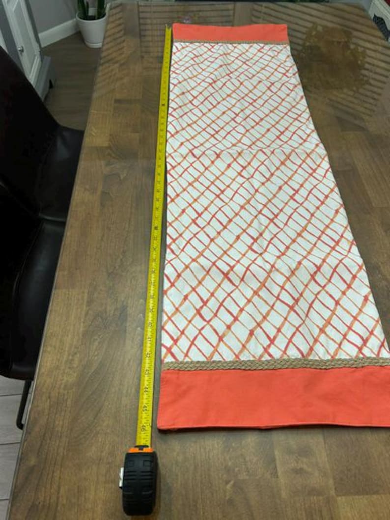 Puede incluir: Un camino de mesa rectangular con un centro blanco de patr&oacute;n de cuadr&iacute;cula naranja, bordeado por extremos de color naranja s&oacute;lido y ribete de yute trenzado. Una cinta m&eacute;trica amarilla al lado muestra una longitud de unos 117 cm.