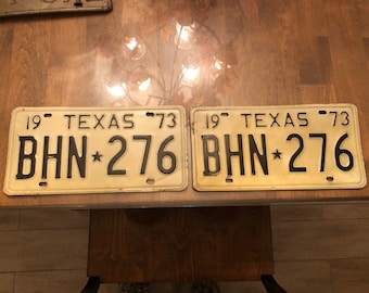 May include: Two vintage Texas license plates from 1973 with the same number "BHN*276" on a wooden surface.