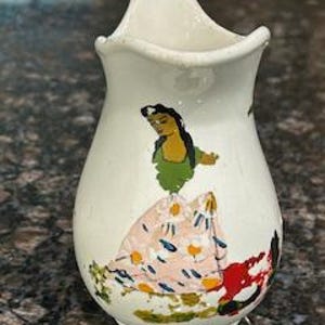 May include: A small, white ceramic vase with a painted design of a woman in a colorful dress. The vase has a curved shape and a scalloped top. The woman's dress is pink with white and yellow accents, and she has a green top.
