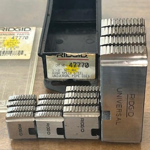 May include: A set of Ridgid universal pipe dies, including a black storage case and several metal dies. The dies are labeled with the Ridgid brand and product information, including "47770" and "High Speed Steel."
