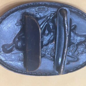 May include: Oval-shaped, dark blue metal belt buckle with a detailed relief of a horse and rider. The buckle has a classic design with a sturdy bar and a hook for securing the belt. The metal has a slightly aged appearance.