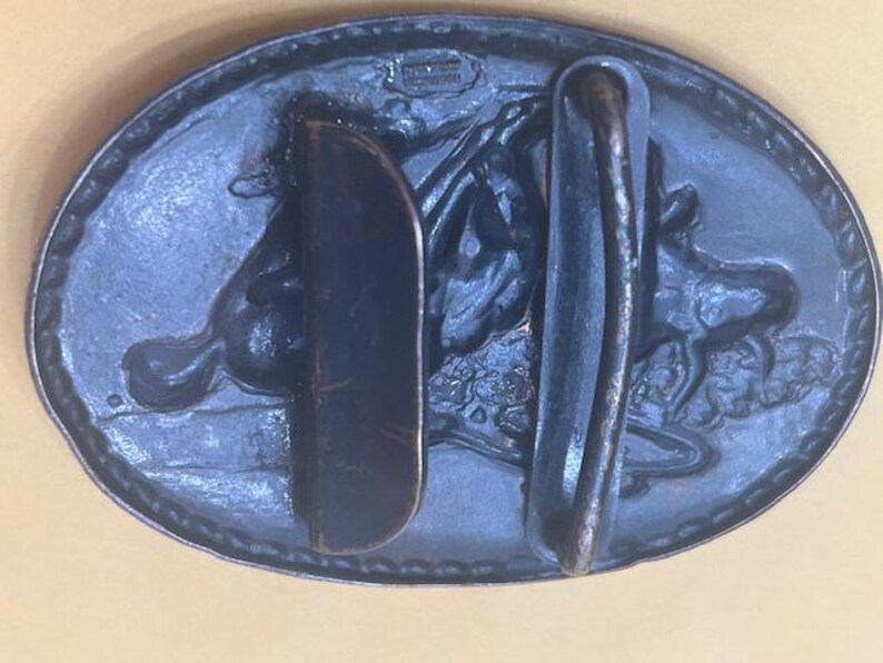 May include: Oval-shaped, dark blue metal belt buckle with a detailed relief of a horse and rider. The buckle has a classic design with a sturdy bar and a hook for securing the belt. The metal has a slightly aged appearance.