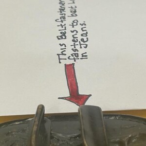 May include: Close-up of a bronze-colored belt buckle with a textured surface. A handwritten note above the buckle reads, "This Belt fastener fastens to belt loop in Jeans."
