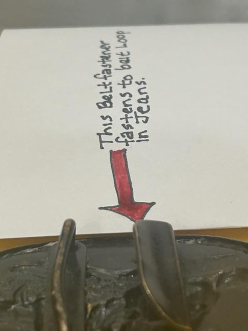May include: Close-up of a bronze-colored belt buckle with a textured surface. A handwritten note above the buckle reads, "This Belt fastener fastens to belt loop in Jeans."