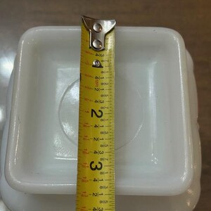 May include: A square white milk glass base with rounded corners, positioned beneath a yellow measuring tape that indicates a width of approximately 4 inches across the center of the object's flat, recessed square surface.