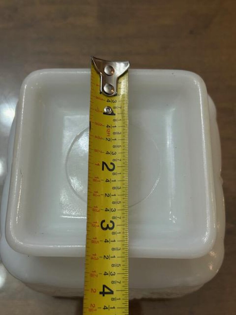 May include: A square white milk glass base with rounded corners, positioned beneath a yellow measuring tape that indicates a width of approximately 4 inches across the center of the object's flat, recessed square surface.