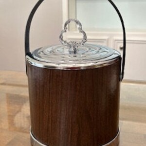 May include: A brown and silver ice bucket with a lid and a handle. The bucket has a wood-grain pattern and a silver band around the bottom.
