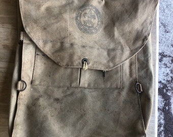 May include: A vintage tan canvas backpack with a Boy Scouts of America logo. The backpack has a flap with a buckle closure and a large pocket on the front.