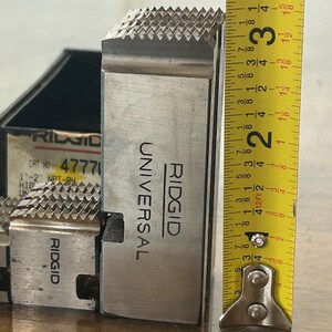 May include: A close-up of a silver-colored RIDGID Universal die head with a toothed cutting surface. The die head is approximately 3 inches tall, as measured by a yellow tape measure. The product is likely for plumbing or pipe fitting.