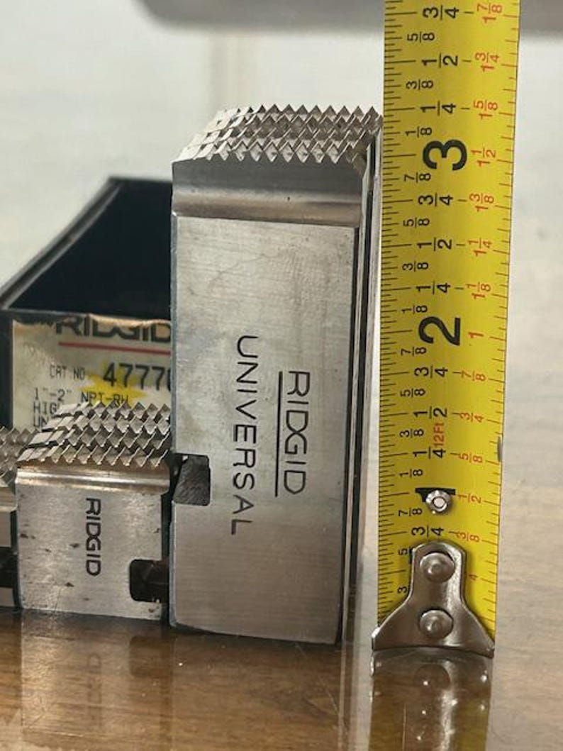 May include: A close-up of a silver-colored RIDGID Universal die head with a toothed cutting surface. The die head is approximately 3 inches tall, as measured by a yellow tape measure. The product is likely for plumbing or pipe fitting.