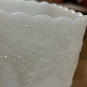 May include: A white milk glass bowl featuring a scalloped rim and a raised relief pattern of grape clusters and vine leaves. The translucent glass surface displays a smooth texture with detailed botanical motifs embossed around the exterior.