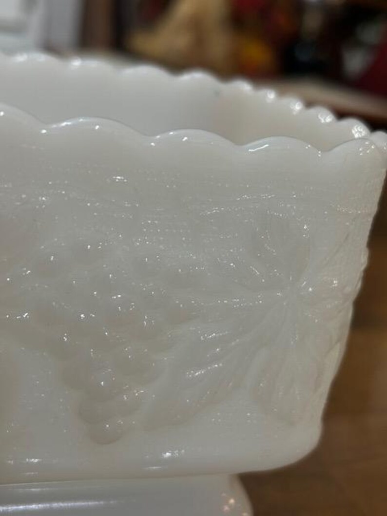 May include: A white milk glass bowl featuring a scalloped rim and a raised relief pattern of grape clusters and vine leaves. The translucent glass surface displays a smooth texture with detailed botanical motifs embossed around the exterior.