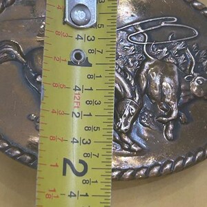 May include: Oval-shaped, bronze-colored belt buckle featuring a cowboy riding a bucking bronco. The buckle is approximately 2 inches (5 cm) in length and has a rope-like border. The design is detailed and shows the cowboy and horse in action.