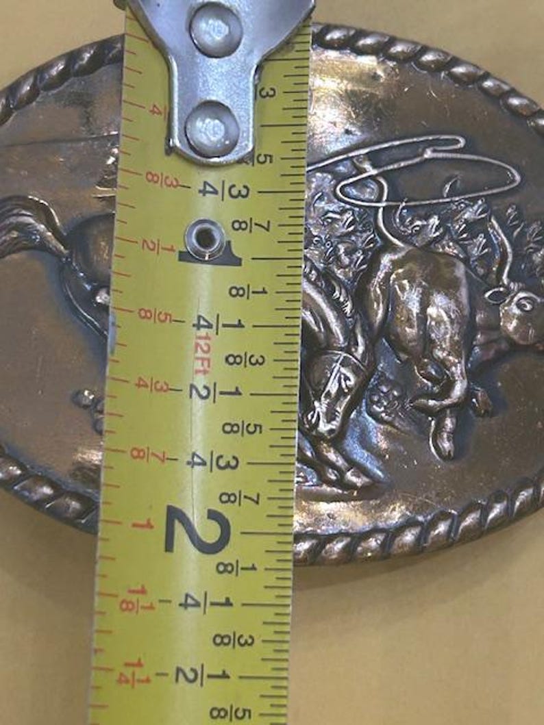 May include: Oval-shaped, bronze-colored belt buckle featuring a cowboy riding a bucking bronco. The buckle is approximately 2 inches (5 cm) in length and has a rope-like border. The design is detailed and shows the cowboy and horse in action.