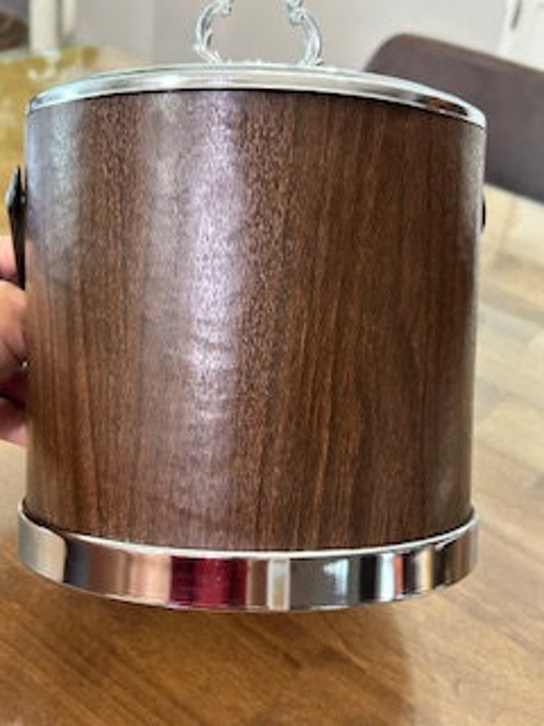 May include: A brown wooden ice bucket with a silver lid and handles. The bucket has a silver band around the top and bottom.