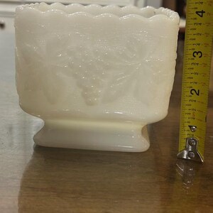 May include: A rectangular white milk glass planter featuring a raised grape and leaf pattern with a scalloped rim. A yellow measuring tape is positioned vertically next to the object, indicating a height of approximately 4.5 inches.