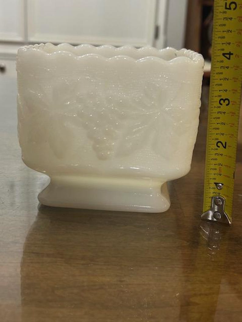 May include: A rectangular white milk glass planter featuring a raised grape and leaf pattern with a scalloped rim. A yellow measuring tape is positioned vertically next to the object, indicating a height of approximately 4.5 inches.