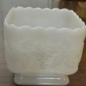May include: A square white milk glass planter featuring a scalloped top edge and a raised relief pattern of grape clusters and leaves on all sides. The vessel sits on a small, integrated square base with a smooth, opaque finish.
