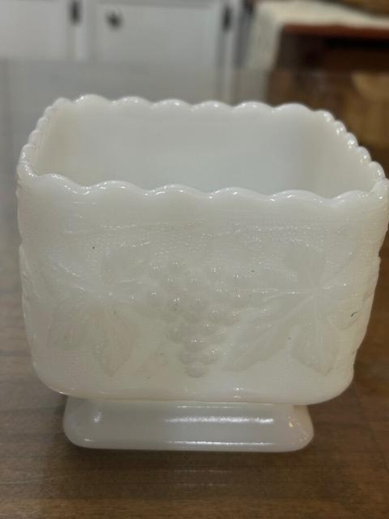May include: A square white milk glass planter featuring a scalloped top edge and a raised relief pattern of grape clusters and leaves on all sides. The vessel sits on a small, integrated square base with a smooth, opaque finish.