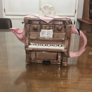 May include: A pink and brown ceramic teapot shaped like an old-fashioned organ with a musical score on the front. The teapot has a pink handle and spout.