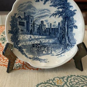 May include: A round ceramic saucer with a scalloped edge featuring a blue and white transferware pattern. The design depicts a large stone manor house, a horse-drawn carriage with a driver, and a large tree under a cloudy sky on a cream base.
