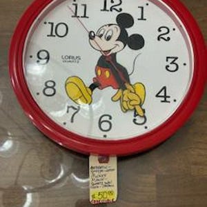 May include: Red circular wall clock with a white face featuring Mickey Mouse in a red and yellow outfit. The clock has black numbers and a black hand.