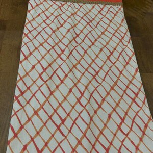 Puede incluir: Un camino de mesa rectangular de tela con fondo blanco y un patr&oacute;n de celos&iacute;a de diamantes en naranja y rojo. El borde superior tiene una banda naranja s&oacute;lida separada del patr&oacute;n principal por un borde trenzado fino de color beige.