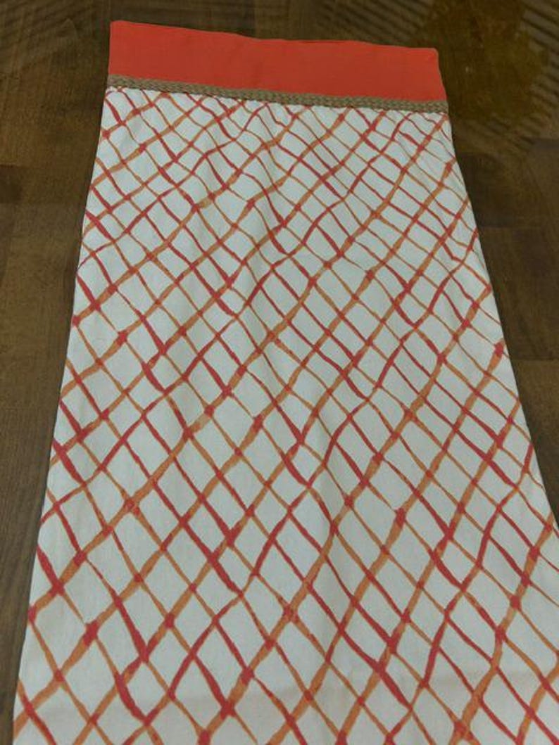 Puede incluir: Un camino de mesa rectangular de tela con fondo blanco y un patr&oacute;n de celos&iacute;a de diamantes en naranja y rojo. El borde superior tiene una banda naranja s&oacute;lida separada del patr&oacute;n principal por un borde trenzado fino de color beige.