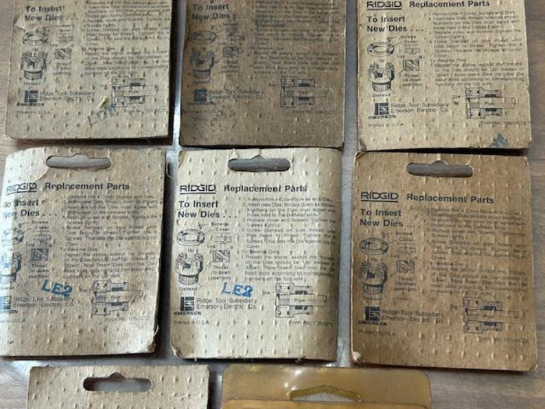 May include: A collection of vintage RIDGID replacement parts packaging. The cardboard packaging is tan with black text and illustrations. The text includes "To Insert New Dies" and "LE2". The packaging is for Ridge Tool Subsidiary Emerson Electric Co.