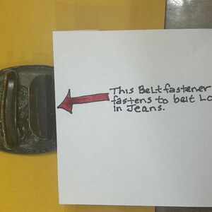 May include: Oval-shaped, dark metal belt buckle with a raised design. A handwritten note indicates the buckle fastens to a belt loop in jeans. The buckle is on a yellow surface.