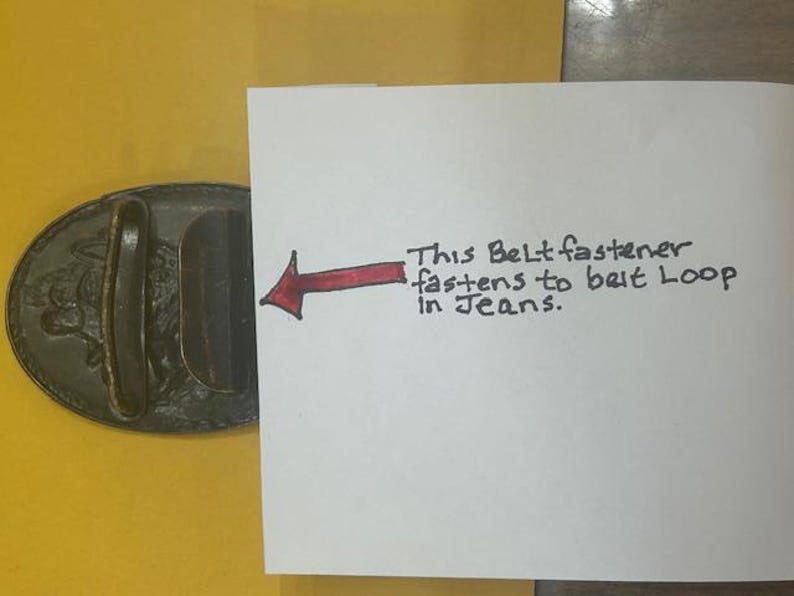 May include: Oval-shaped, dark metal belt buckle with a raised design. A handwritten note indicates the buckle fastens to a belt loop in jeans. The buckle is on a yellow surface.