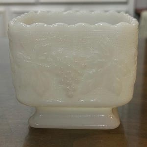 May include: A square milk glass planter featuring a raised relief pattern of grape clusters and leaves on all sides. The vessel has a scalloped top edge and sits on a raised rectangular base, showcasing a smooth, opaque white finish throughout.