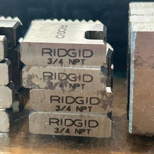 May include: Close-up of several metal RIDGID 3/4 NPT die heads. The die heads are stacked and have the RIDGID logo and size information printed on them. The metal has a slightly worn appearance.