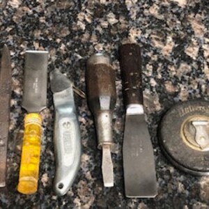 May include: A collection of vintage tools including a putty knife, a utility knife, a screwdriver, a tape measure, and a file. The tools are all in various states of wear and tear, suggesting they have been used extensively.