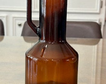 May include: A brown glass bottle with a handle and a cork stopper. The bottle has a rounded bottom and a narrow neck.