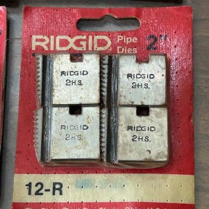 May include: RIDGID 2" pipe dies in a red and tan package. The package has the text "RIDGID Pipe Dies" and "2" printed on it. The dies are silver and have the text "RIDGID 2H.S." printed on them. The package also has the text "12-R" printed on it.