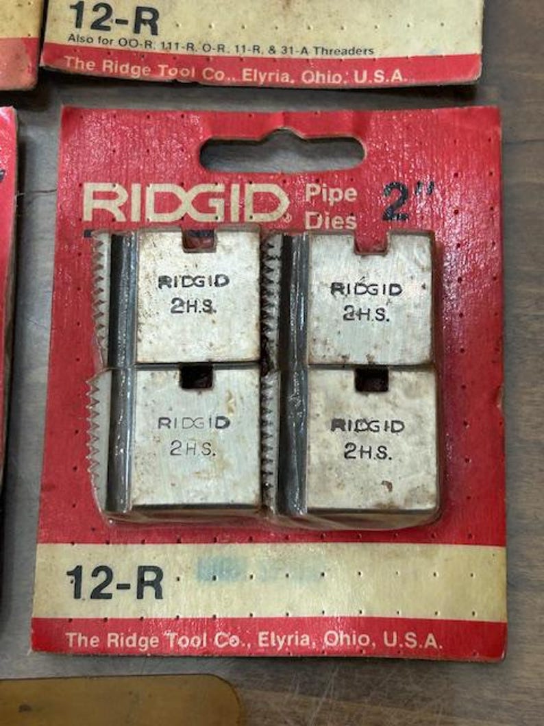 May include: RIDGID 2" pipe dies in a red and tan package. The package has the text "RIDGID Pipe Dies" and "2" printed on it. The dies are silver and have the text "RIDGID 2H.S." printed on them. The package also has the text "12-R" printed on it.