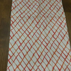 Puede incluir: Un camino de mesa rectangular de tela con fondo blanco y un patr&oacute;n de cuadr&iacute;cula diagonal repetitivo. Las l&iacute;neas de la cuadr&iacute;cula est&aacute;n representadas en varios tonos de naranja y coral, creando un efecto pintado a mano sobre el tejido.