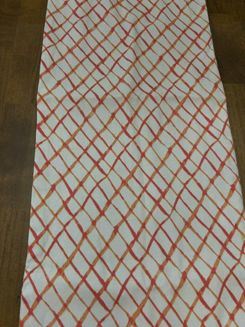 Puede incluir: Un camino de mesa rectangular de tela con fondo blanco y un patr&oacute;n de cuadr&iacute;cula diagonal repetitivo. Las l&iacute;neas de la cuadr&iacute;cula est&aacute;n representadas en varios tonos de naranja y coral, creando un efecto pintado a mano sobre el tejido.