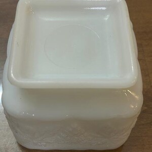 May include: A square white milk glass container featuring a raised embossed pattern of leaves and berries along the sides. The object has a smooth, flat square base and a slightly recessed center on the bottom surface, with a glossy finish.