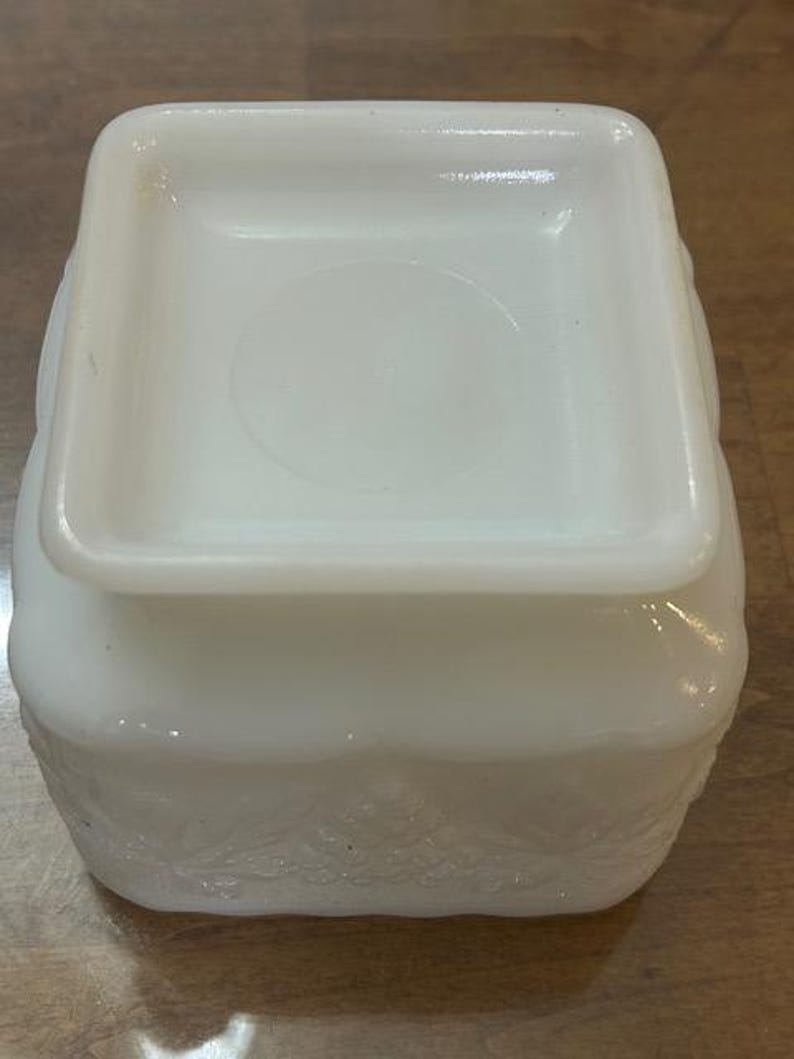 May include: A square white milk glass container featuring a raised embossed pattern of leaves and berries along the sides. The object has a smooth, flat square base and a slightly recessed center on the bottom surface, with a glossy finish.