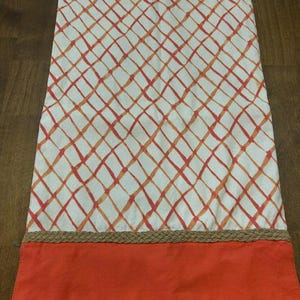 Puede incluir: Un camino de mesa rectangular con base de tela blanca y un patr&oacute;n de cuadr&iacute;cula de diamantes en naranja y rojo. El borde inferior est&aacute; rematado con un ribete de yute trenzado en color tostado y una franja de tela naranja s&oacute;lida.