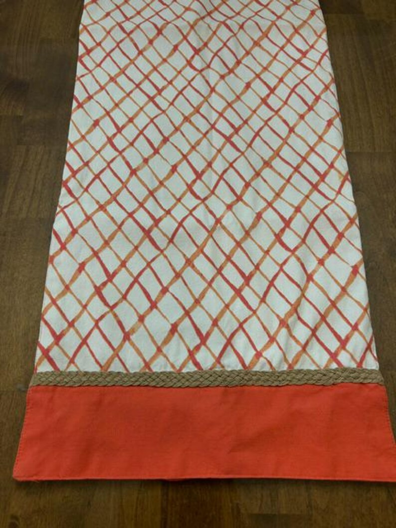 Puede incluir: Un camino de mesa rectangular con base de tela blanca y un patr&oacute;n de cuadr&iacute;cula de diamantes en naranja y rojo. El borde inferior est&aacute; rematado con un ribete de yute trenzado en color tostado y una franja de tela naranja s&oacute;lida.