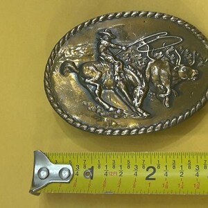 May include: Oval-shaped, vintage metal belt buckle featuring a cowboy on a bucking bronco roping a bull. The buckle has a detailed relief design with a rope-like border. The buckle measures approximately 3 inches in length.
