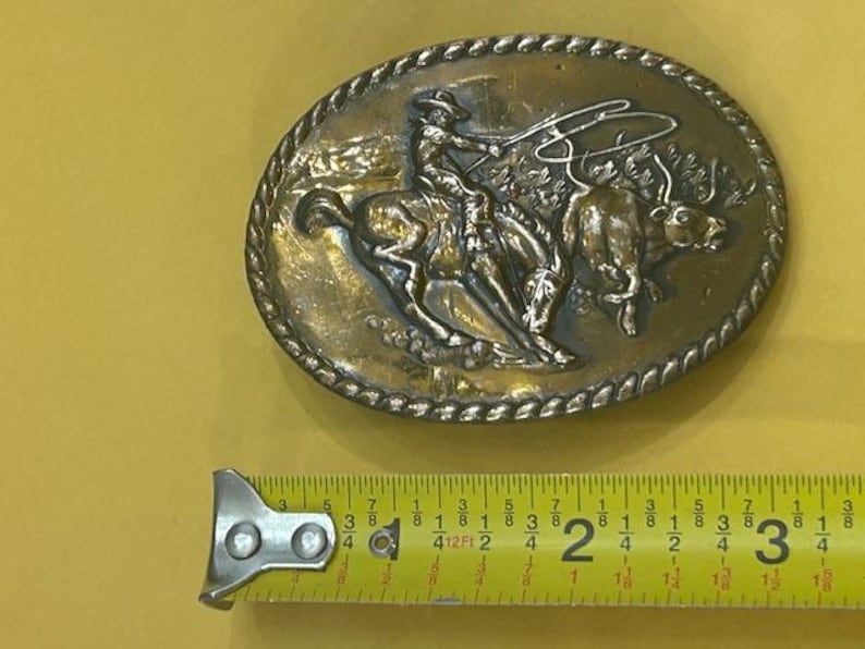 May include: Oval-shaped, vintage metal belt buckle featuring a cowboy on a bucking bronco roping a bull. The buckle has a detailed relief design with a rope-like border. The buckle measures approximately 3 inches in length.