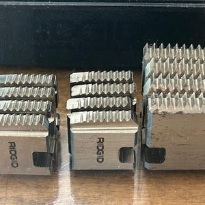 May include: Three silver-colored RIDGID pipe threading dies of varying sizes. Each die has a series of sharp, angled teeth for cutting threads. The RIDGID logo is visible on two of the dies. The dies are arranged on a light brown surface.
