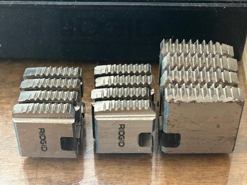 May include: Three silver-colored RIDGID pipe threading dies of varying sizes. Each die has a series of sharp, angled teeth for cutting threads. The RIDGID logo is visible on two of the dies. The dies are arranged on a light brown surface.