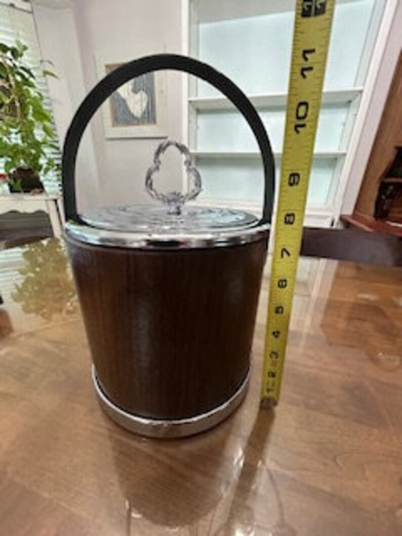 May include: A brown wooden ice bucket with a chrome trim and a silver lid with a crystal handle. The bucket is 9 inches tall.